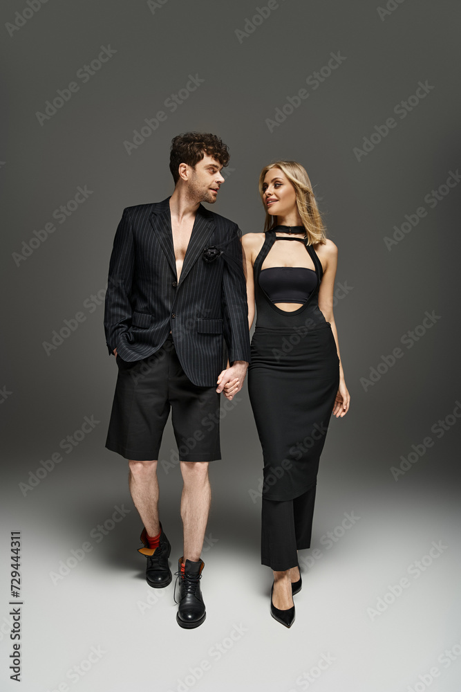 full length of couple in trendy black attire holding hands while standing on dark grey background full length of couple in trendy black attire holding hands while standing on dark grey background