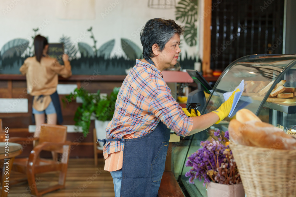 Businessman shop owner elderly Asian retired age pensioner employee cafe small business using blue cleaning cloth clean  glass cabinet bakery after cafe closed intently smiling corners of his mouth