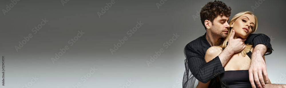 Fototapeta premium handsome curly man in pinstripe blazer embracing attractive blonde woman on grey background , banner