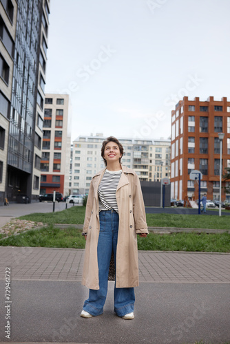 Full length outdoor portrait of happy owner of new apartment in comfortable residential area, standing among multi-storey buildings, admiring surroundings, looking up with happy facial expression