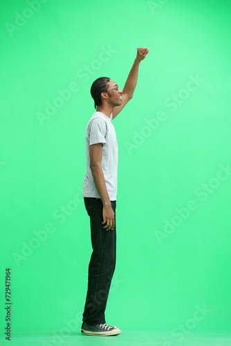 A man in a gray T-shirt, on a green background, in full height, raised his hand up © Katsiaryna