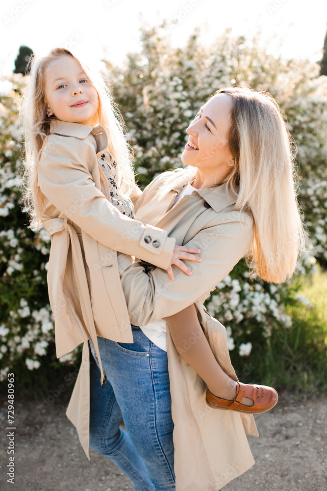 Fototapeta premium Happy young mother holding child girl 4-5 year old wearing beige similar jacket in sun light outdoor. Motherhood. Spring season.