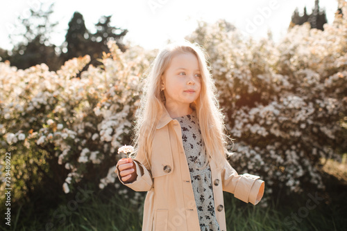 Cute blonde baby girl 4-5 year old wearing beige jacket with flowers at background outdoor. Spring season. Childhood.