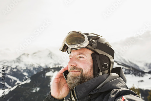 Portrait of happy man while up on ski slope, wearing helmet and goggles, talking on his cell phone.. Zillertal, Austria photo