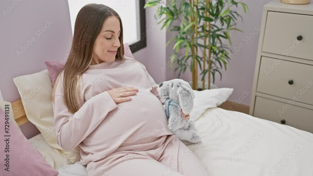 A young pregnant hispanic woman in pink pajamas holds a plush toy while relaxing on a bed in a well-lit bedroom at home. A young pregnant hispanic woman in pink pajamas holds a plush toy while relaxing on a bed in a well-lit bedroom at home.