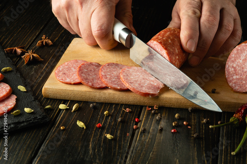 A knife in the hand of a chef for cutting salami sausage on a kitchen cutting board. Idea for serving a sausage dish