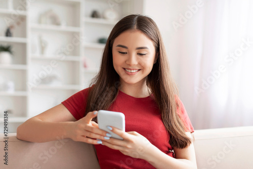 Smiling young asian woman focused on texting with her smartphone at home