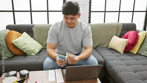 Asian man counting hong kong dollars in a modern living room with laptop and colorful cushions photo