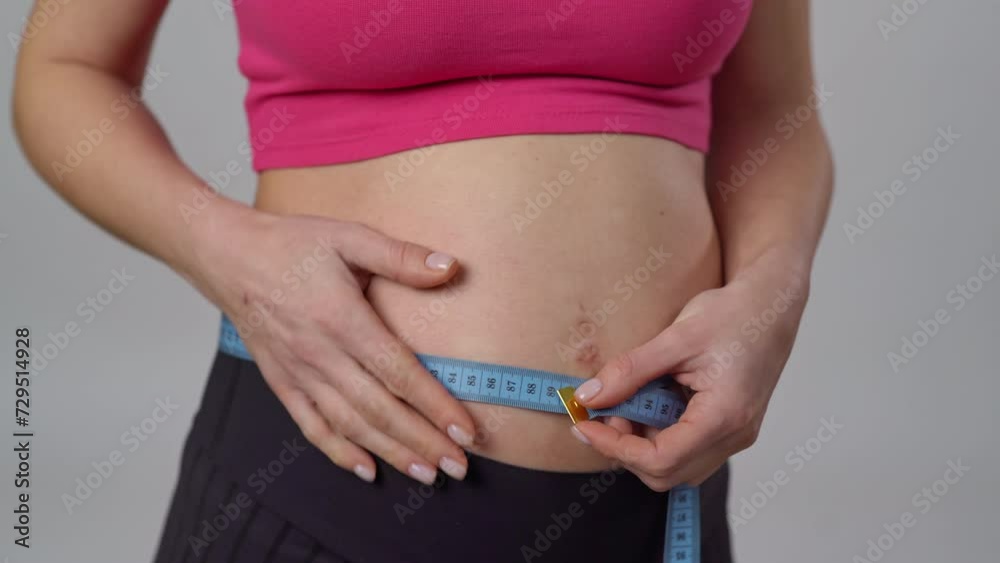 Close-up. Unrecognizable pregnant woman measuring her belly with a measuring tape while standing on a white background. A pregnant woman gently strokes her belly