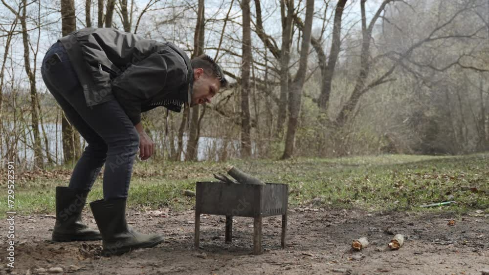Man blows his breath into fire in metal barbecue in forest. Guy blows sharply on smoking coals and firewood, causing increase in temperature and heat. Making fire in nature, survival skills. Man blows his breath into fire in metal barbecue in forest. Guy blows sharply on smoking coals and firewood, causing increase in temperature and heat. Making fire in nature, survival skills.