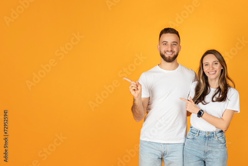 A joyful man and woman in white t-shirts and blue jeans point to their side with smiling faces © Prostock-studio
