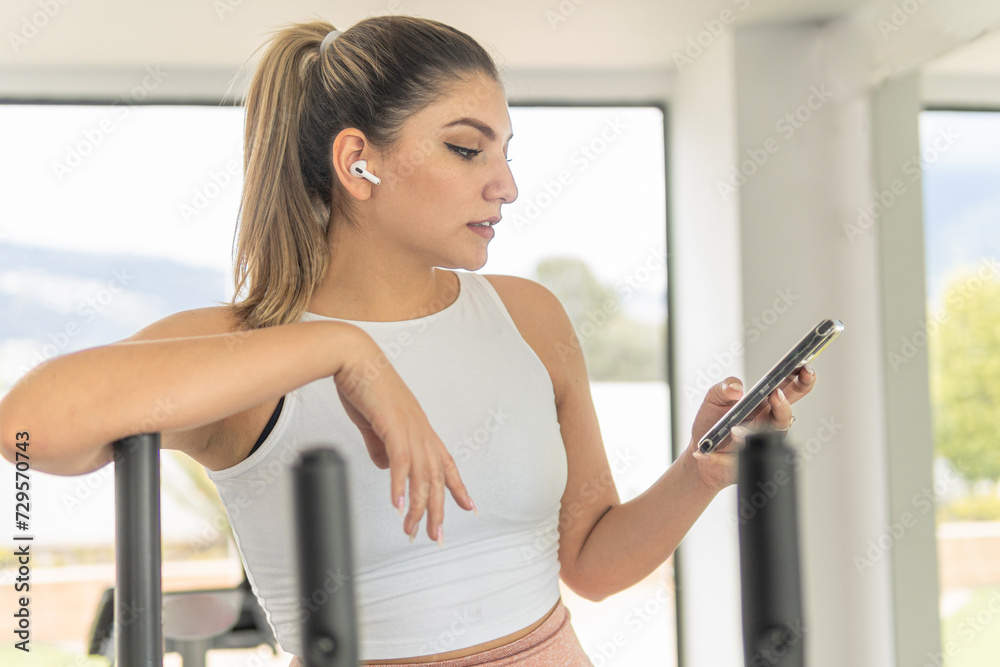 young woman with sports top choosing music on her cell phone to listen to it with her wireless headphones at the gym