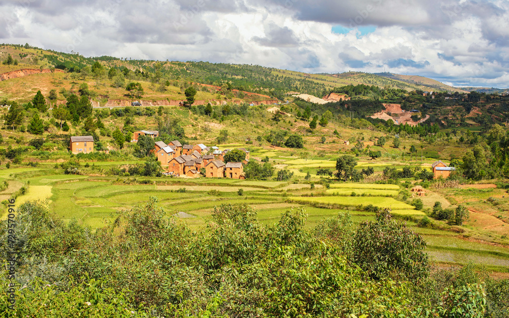 Tsiafahy, Madagascar - April 25, 2019: Typical Madagascar landscape in region near Tsiafahy, small hills covered with green grass and bushes, red clay houses and rice fields near