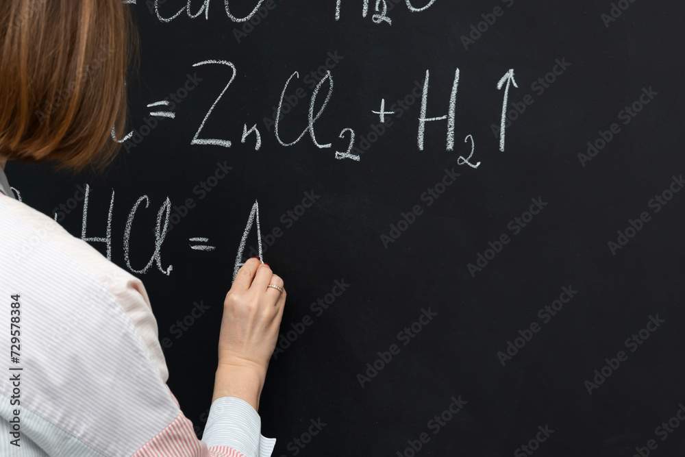Back view of a female student writing the formula of a chemical reaction on the blackboard Back view of a female student writing the formula of a chemical reaction on the blackboard