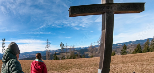 Couple at summit cross on alpine meadow in West Styrian highland in Voitsberg, Styria, Austria. Hiking in remote landscape of soft hills in spring. Scenic view of idyllic mountains of Lavanttal Alps photo