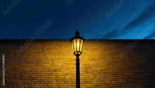 Um poste de iluminação pública com luminária vintage, com um muro de tijolos laranjados, iluminado ao fundo. Foto noturna. photo