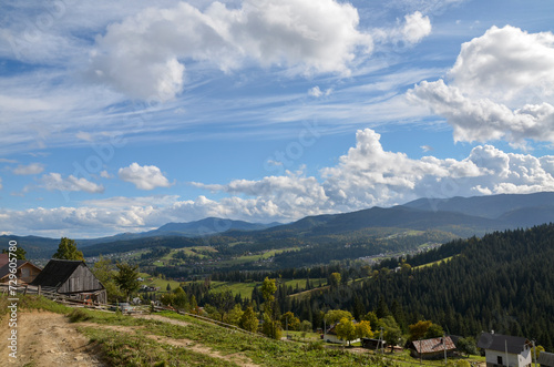 View of a village with wooden houses in a mountainous area surrounded by green meadows and mountains in the distance covered with dense forest under a clear sky with white clouds. Carpathians, Ukraine