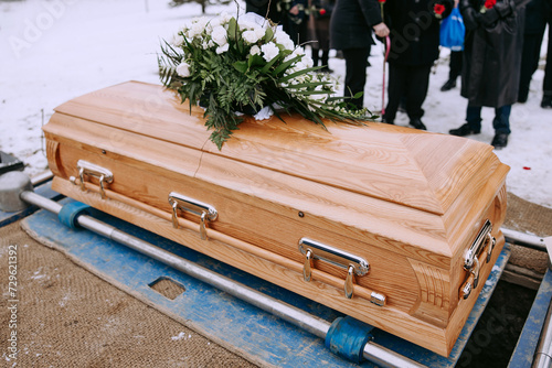 Canvas-taulu A wooden coffin with a bouquet of white flowers on the lid stands over a dug grave at the cemetery