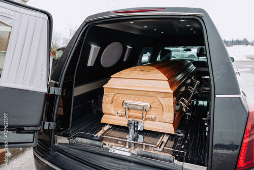A coffin made of light wood stands in the trunk of a black hearse. Funeral and farewell ceremony. Closeup photo of a funeral casket photo