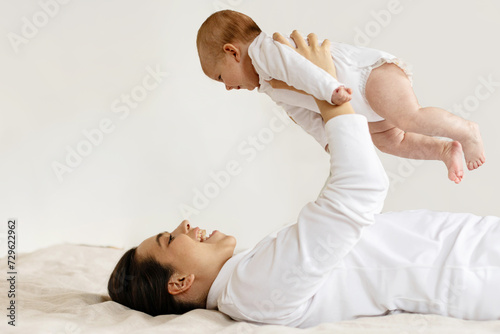 Happy motherhood. Mother playing with baby daughter and raising kid up while lying on bed, mom cuddling and having fun with child, side view
