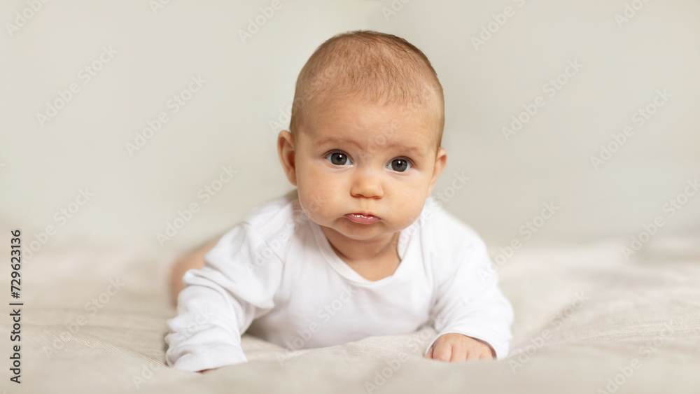 Baby care. Infant child relaxing on bed at home, little boy or girl lying on his tummy in bedroom, looking at camera, copy space