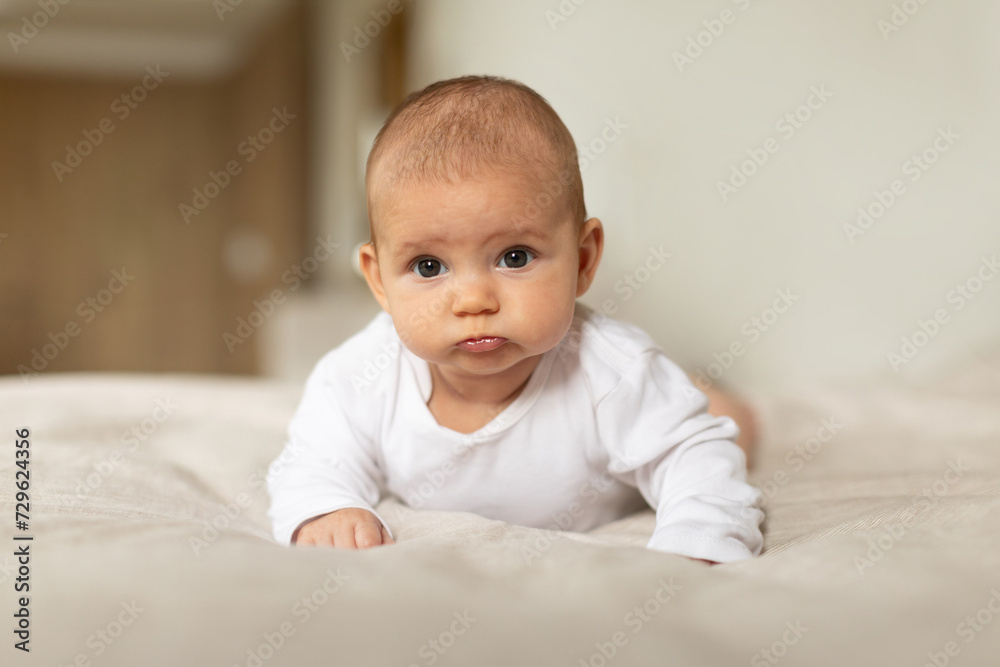 Childcare concept. Little baby girl lying on tummy on bed at home, cute infant in white bodysuit in bedroom, looking at camera, copy space