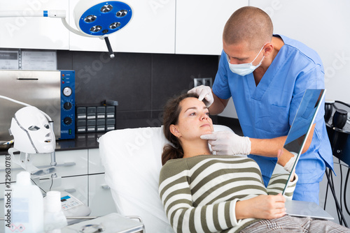 Young woman looking at mirror while professional cosmetician explaining her future hardware facial procedure in medical esthetic office..