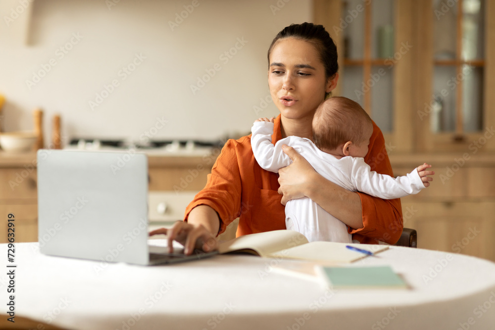 Freelance and motherhood. Young mother working remotely on laptop and taking care about baby, holding infant daughter and using computer in kitchen Freelance and motherhood. Young mother working remotely on laptop and taking care about baby, holding infant daughter and using computer in kitchen
