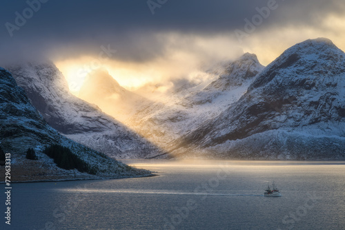 Snowy mountains in low clouds, bright sunbeams, boat in sea bay at colorful sunset in winter. Lofoten islands, Norway. Amazing landscape with rocks in snow, golden sun rays. North seaside. Nature
