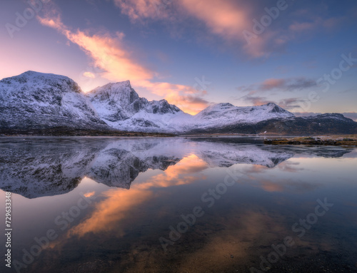 Beautiful snowy mountains and colorful sky with clouds at sunset in winter in Lofoten islands, Norway. Landscape with rocks in snow, sea coast, reflection in water at dusk, purple sky with pink clouds