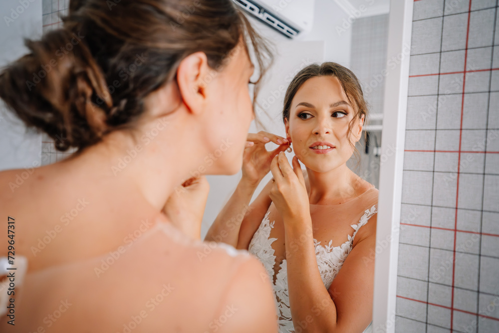 Valmiera, Latvia - July 7, 2023 - A bride in a lace dress is putting on her earring while looking at her reflection in a mirror. Valmiera, Latvia - July 7, 2023 - A bride in a lace dress is putting on her earring while looking at her reflection in a mirror.