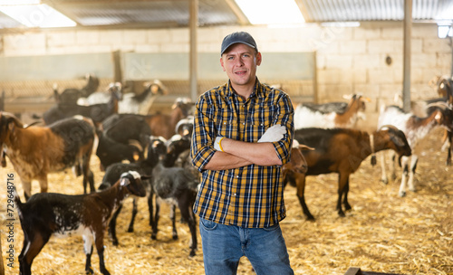 Confident positive male farmer, engaged in breeding dairy goats, posing in goat shed .. photo