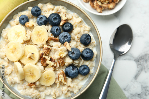 Tasty oatmeal with banana, blueberries, walnuts and milk served in bowl on white marble table, flat lay