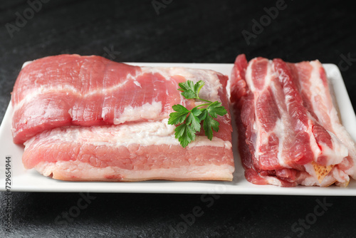Pieces of raw pork belly and parsley on black textured table, closeup
