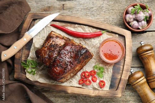 Piece of baked pork belly served with sauce and chili pepper on wooden table, flat lay