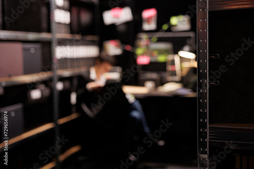 Selective focus of shelves full with evidence files, in background private investigator reading crime scene report. Police officer working late at night at missing person case in arhive room. photo