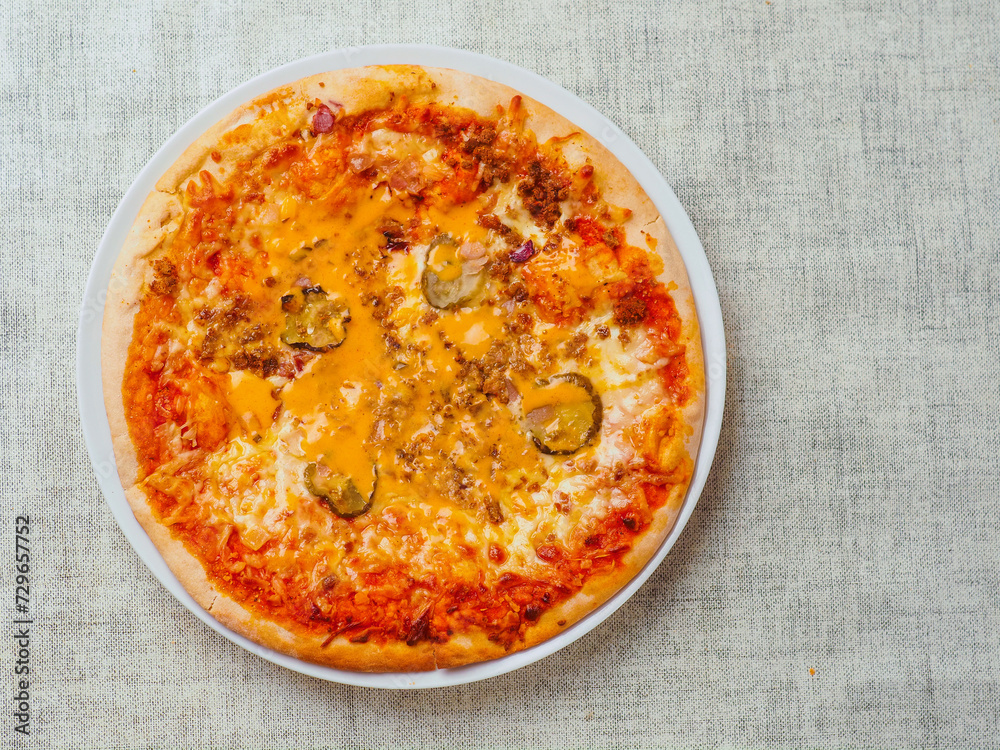 Cooked burger style Italian pizza with tomato sauce, meat, cucumber and cheese on round metal tray and light color table cloth. Ready meals product for cooking home. High quality ingredients Cooked burger style Italian pizza with tomato sauce, meat, cucumber and cheese on round metal tray and light color table cloth. Ready meals product for cooking home. High quality ingredients