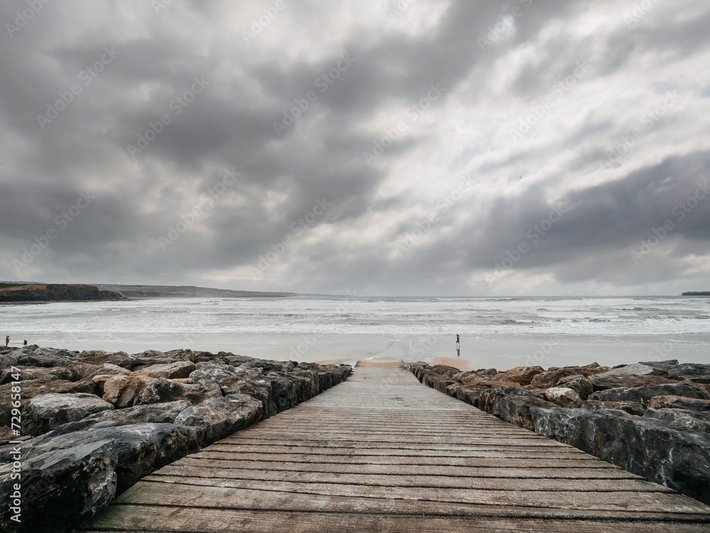 Ramp to a vast sandy beach and rough ocean water, silhouette of a person walking along shows the scale of the place. Lahinch, county Clare, Ireland. Dark dramatic sky. Rough nature scene.