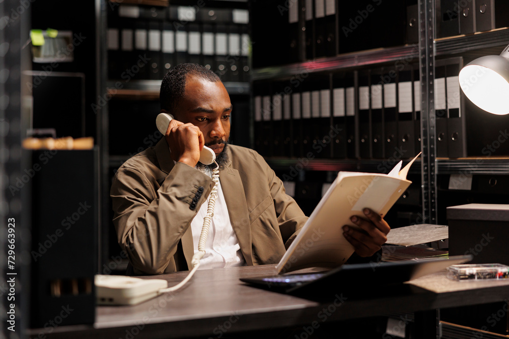 Private detective discussing investigation with client on landline phone. African american investigator reading crime case file and talking on telephone with law enforcement professional Private detective discussing investigation with client on landline phone. African american investigator reading crime case file and talking on telephone with law enforcement professional