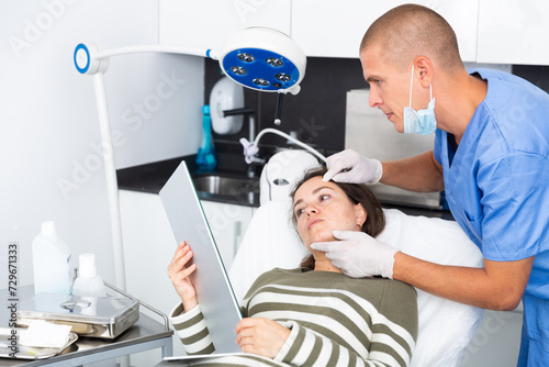 Portrait of woman sitting on couch and looking at mirror during appointment with doctor at aesthetic clinic