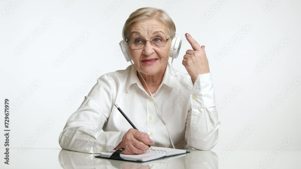 Romantic neat old lady with blonde hair white shirt sitting at table, listening to music, making notes with pencil, waving index finger, white background. High quality 4k footage Romantic neat old lady with blonde hair white shirt sitting at table, listening to music, making notes with pencil, waving index finger, white background. High quality 4k footage