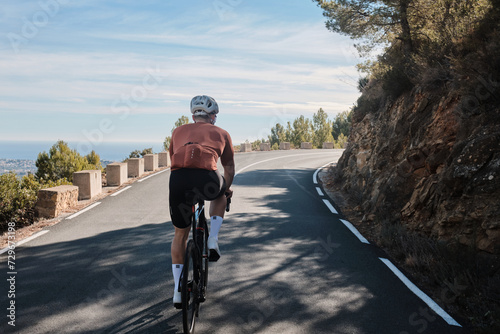 Man cyclist in full cycling kit riding a road bicycle on the mountain road Bernia, Costa Blanca, Alicante, Spain. Male professional cyclist climbing in the spanish mountains back view. photo
