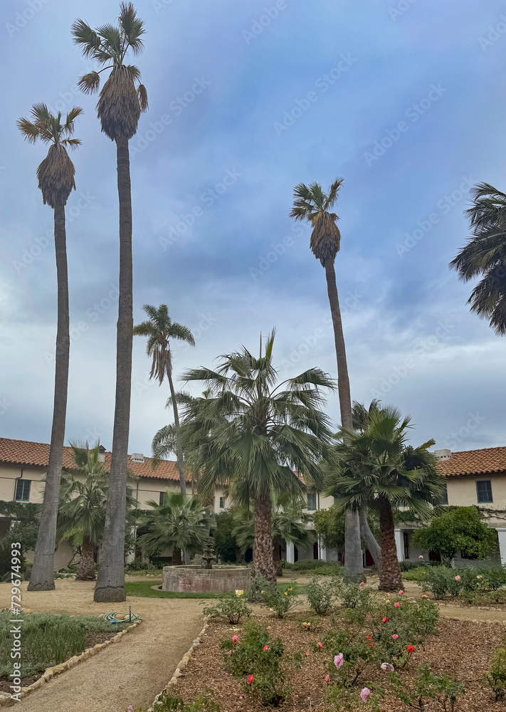 Santa Barbara, CA, USA - December 18, 2023: Inner courtyard of Old Mission church with tall palm trees, fountain in center, and flowers, all under blue cloudscape