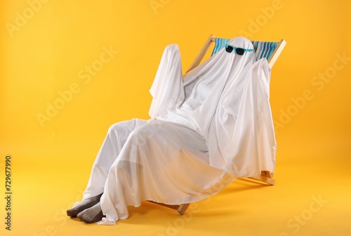 Person in ghost costume and sunglasses relaxing on deckchair against yellow background