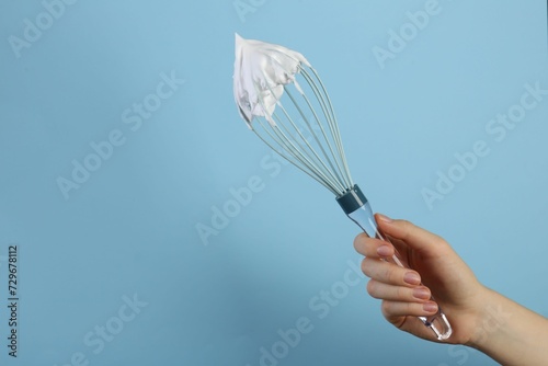 Woman holding whisk with whipped cream on light blue background  closeup. Space for text