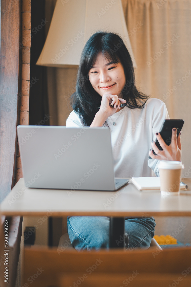 Business freelance concept, A young, smiling Asian woman with a laptop and smartphone, comfortably working in a cozy cafe setting. Business freelance concept, A young, smiling Asian woman with a laptop and smartphone, comfortably working in a cozy cafe setting.