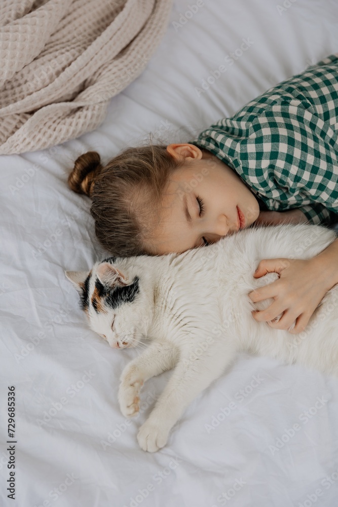 A little blond girl wearing pajamas hugs a domestic cat in her bedroom. The concept of a cozy home morning A little blond girl wearing pajamas hugs a domestic cat in her bedroom. The concept of a cozy home morning