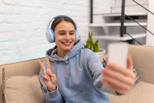 Woman with headphones making selfie on couch showing v sign in home interior.