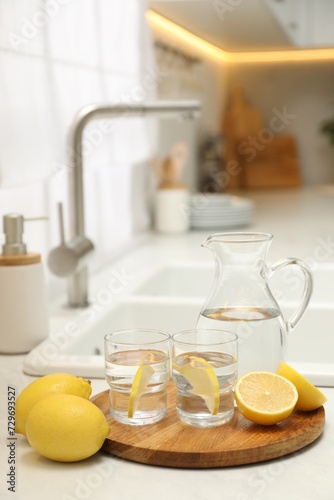 Jug, glasses with clear water and lemons on white table in kitchen