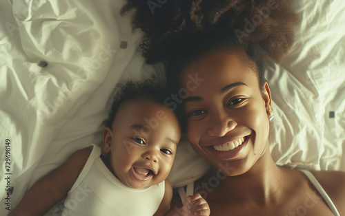 A young African American mother and her baby are lying on a bed together. They are both smiling and looking at the camera